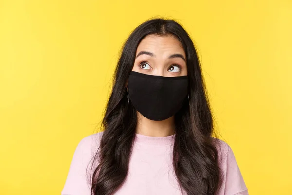 Covid-19, stile di vita social-distanziamento, prevenire il concetto di diffusione del virus. Incuriosito e premuroso ragazza asiatica in maschera, guardando in alto a sinistra pensiero o imaging qualcosa, sfondo giallo — Foto Stock