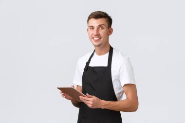 Küçük işletme, kafe ve kafe çalışanları konsepti. Yakışıklı garson, önlük giyen barista, sipariş alıyor, dijital tabletli masaları kurtarıyor, kameraya gülümsüyor, beyaz arka plan
