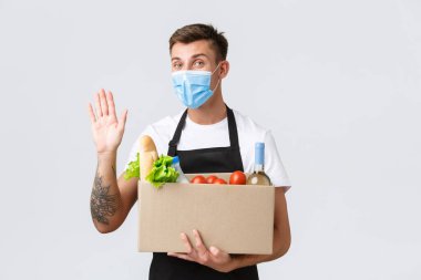 Covid-19, temassız alışveriş ve market teslimatı konsepti. Dost canlısı pazarlamacı yiyecek kutusunu tutuyor, sipariş veriyor, tıbbi maske takarak merhaba demek için el sallıyor, beyaz arka plan...