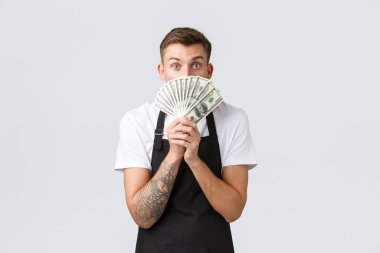 Küçük perakende işi, ödemeler ve çalışanlar konsepti. Heyecanlı ve heyecanlı yakışıklı barista, siyah önlüklü garson, paranın arkasına saklanan, büyük bahşiş ya da ödeme alan, beyaz arka plan