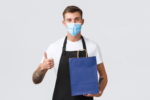 Retail store, shopping during covid-19 and social distancing concept. Cheerful handsome salesman in medical mask and apron provide quality service, show thumbs-up and handing bag with product