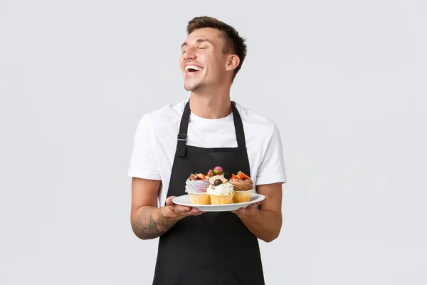 Petit commerce de détail, cafés et concept de boulangerie. Beau barista passionné, vendeur en pâtisserie, les yeux fermés et souriant ravi, assiette avec cupcakes, fond blanc — Photo