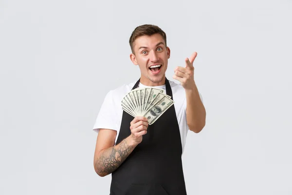 Pequeña empresa minorista, concepto de pagos y empleados. Barista guapo alegre y entusiasta, vendedor invitando a trabajar en la cafetería, sosteniendo el dinero y señalando con el dedo a la cámara — Foto de Stock