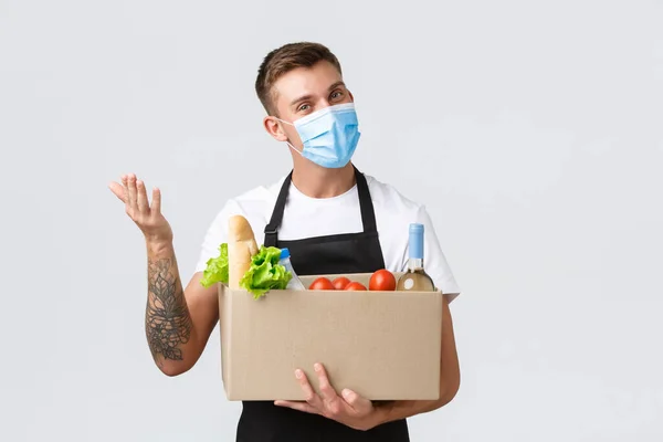 Covid-19, contactless shopping and groceries delivery concept. Friendly salesman hold box with groceries, delivering order, waving hand to say hello wearing medical mask, white background — Stock Photo, Image