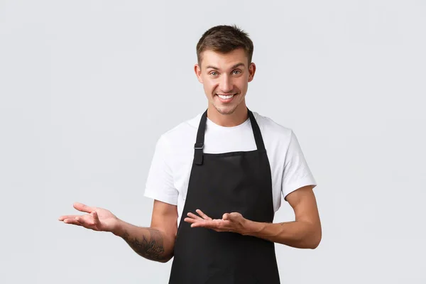 Cafe and restaurants, coffee shop owners, retail concept. Handsome cheeky salesman, waiter in black apron pointing left, inviting come inside, showing way or introducing discounts, white background — Stock Photo, Image