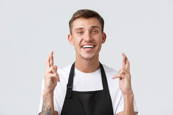 Café och restauranger, caféägare och butikskoncept. Närbild av hoppfull vacker servitör, säljare i svart förkläde kors fingrar lycka och leende optimism, be över vit bakgrund — Stockfoto