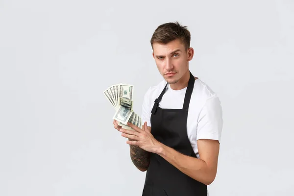 Pequeña empresa minorista, concepto de pagos y empleados. Vendedor guapo descarado, dueño de la cafetería contando ingresos, mostrando dinero y sonriendo satisfecho, de pie fondo blanco — Foto de Stock