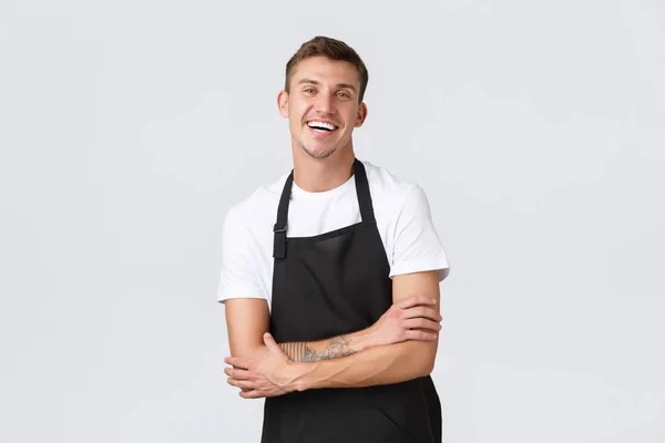 Small business owners, coffee shop and staff concept. Enthusiastic smiling, handsome waiter in black apron, laughing happily, serving orders to clients, working in cafe, standing white background — Stock Photo, Image