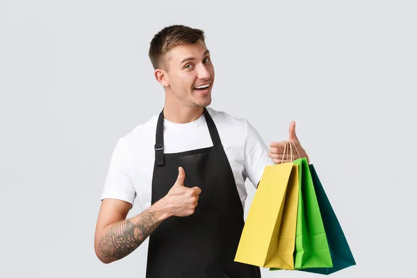 Retail store, shopping and employees concept. Friendly cheerful salesman, showing thumbs-up and products in eco-bags, guarantee best service, handing over purchased items, white background