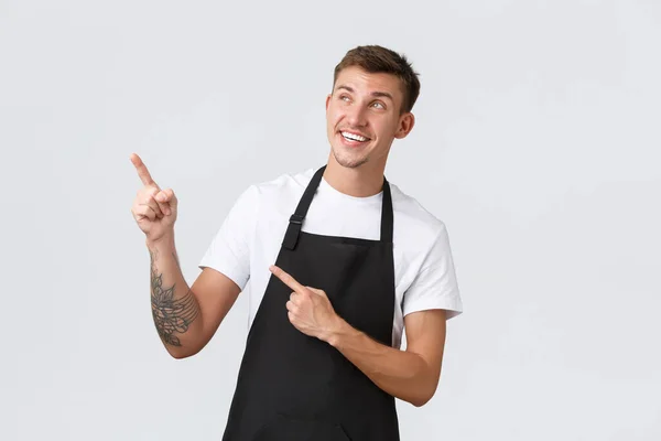 Small business owners, coffee shop and staff concept. Smiling pleased and happy handsome barista in black apron, cafe employee pointing and looking upper left corner satisfied, showing advertisement — Stock Photo, Image