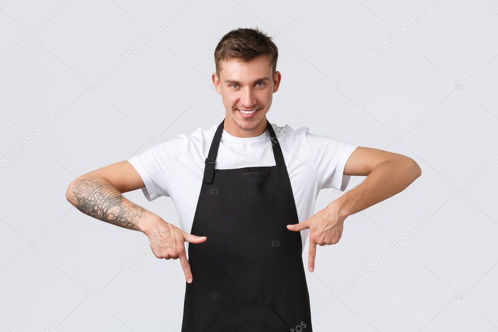 Small business owners, coffee shop and staff concept. Assured handsome blond guy working in cafe, restaurant waiter pointing fingers down and smiling, give guarantee, recommending place enjoy drink