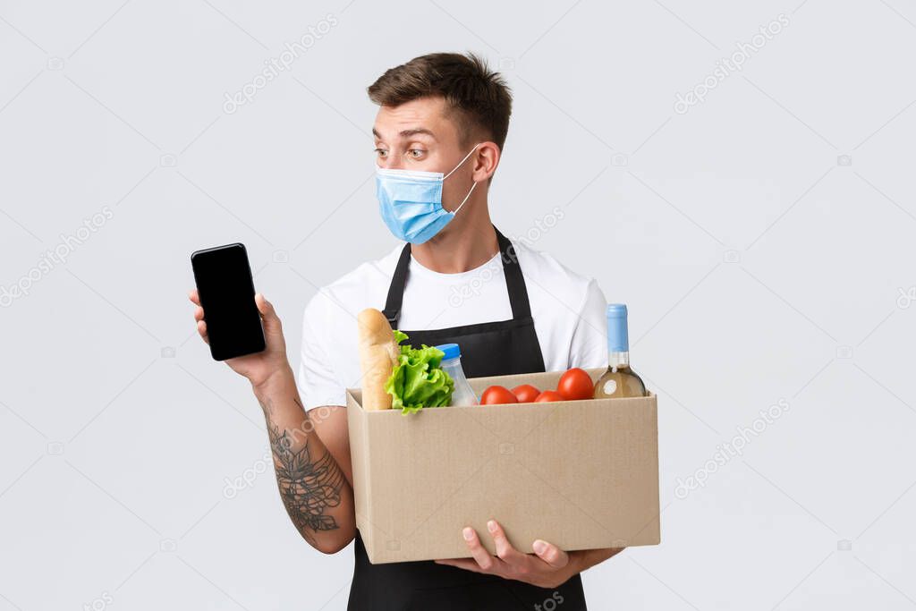 Covid-19, contactless shopping and groceries delivery concept. Handsome salesman in medical mask suggest way of ordering grocery products using smartphone app, show mobile phone and box
