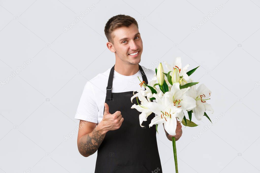 Small business, retail and employees concept. Handsome salesman in black apron, made beautiful flowers, florist in store selling lilies, smiling friendly at customer, showing thumbs-up