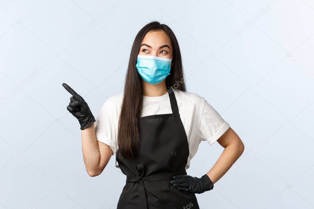 Covid-19 pandemic, social distancing, small business and preventing virus concept. Pleased smiling asian female coffee shop barista in medical mask and gloves seeing good promo at upper left corner