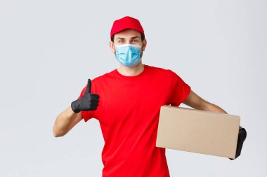 Paketler ve paket teslimatı, karantina teslimatı, nakil emirleri. Kırmızı üniformalı neşeli kurye, eldivenler ve yüz maskesi, baş parmak kaldırma, temassız teslimat önerilir, sipariş ile tutulan kutu