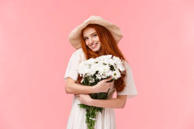 Güzel ve cezbedici romantik sevimli, saman şapkalı aptal kızıl kız, güzel beyaz çiçekleri kucaklıyor, buket hediyesini alıyor ve gülümsüyor kamera küstahça, durgun pembe arka plan