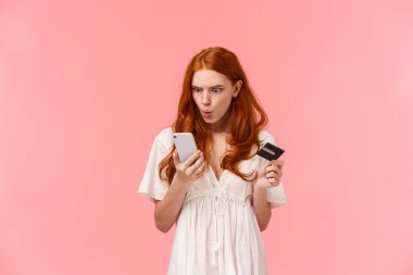 Sinirli ve kızgın, kafası karışmış kızıl saçlı kadın akıllı telefon görüntüsünden memnun değil, bir şeylerin yanlış gittiğinden şikayet ediyor, banka hesabını boş kontrol ediyor, elinde kredi kartı tutuyor.