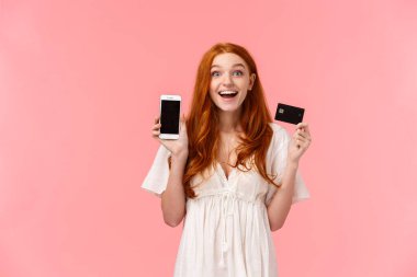 Çekici, keyifli ve büyüleyici mutlu kızıl saçlı kız mutlu ve mutlu, elinde kredi kartıyla, akıllı telefon ekranını göstererek, harika bir alışveriş yaptı, arkadaşına böbürlenerek, pembe arka planda