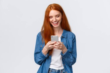 Sohbet, iletişim ve mobil konsept. Neşeli kızıl saçlı, gülümseyen kız mesaj atıyor, gülüyor, komik video bloğu izliyor, akıllı telefon görüntüsü veriyor, mutlu görünüyor, beyaz arka planda duruyor.