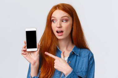İlginç, güzel kızıl saçlı beyaz kadın tatilden fotoğraflar gösteriyor, akıllı telefon ekranını işaret ediyor, beyaz arka plan üzerinde duruyor,