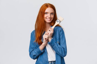 Kot gömlekli, yaratıcı, güzel, kızıl saçlı kadın, yeni sanat eserleri yaratıyor, resim yapıyor, kendi odasında tamirat yapıyor, elinde boya fırçası tutuyor ve gülümsüyor, baştan çıkarıcı, beyaz arka planda duruyor.