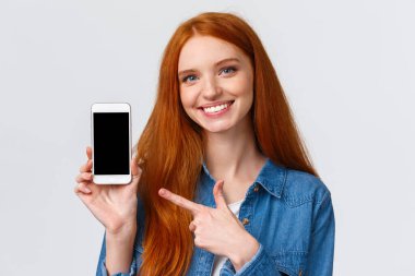 Kız flörtünü bu uygulamada bulmuş. Mavi gözlü neşeli, çekici kızıl kadın tavsiye veriyor, akıllı telefon ekranını gösteriyor ve hareket eden bir ekranı işaret ediyor, gülümsüyor tavsiye veriyor.