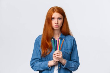 Kasvetli ve kararsız, kararsız, çekingen, çekingen, çekingen kızıl saçlı kız elinde renkli kalemler olan ve kameraya bakan, mütevazı bir ifadeyle, suratını asan.