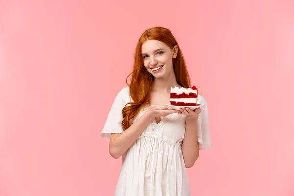 Lovely, romantic and alluring redhead girlfriend baked delicious surprise for valentines day date, holding peace cake on plate and smiling tempting with sassy, coquettish expression, pink background — Stock Photo, Image