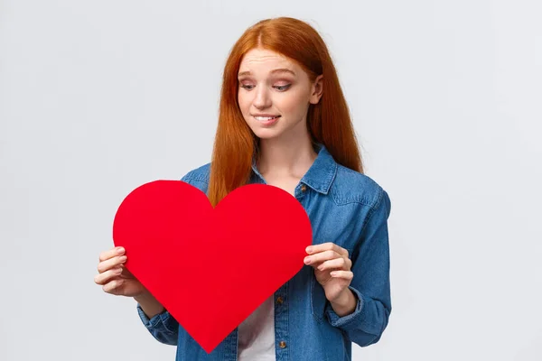 Indecisive cute and timid, shy redhead girl wants confess in love, show sympathy unsure give present on valentines day or not, holding big red heart, biting lip worried, white background