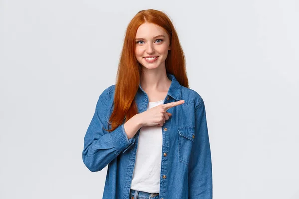 Taille-up portret vriendelijk glimlachend, gelukkig roodharige zelfverzekerde vrouwelijke student in denim shirt, tonen waar goede cursussen online, wijzende vinger rechts en grijnzend, nodigen join, witte achtergrond — Stockfoto