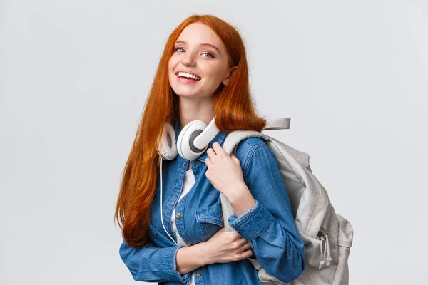 Vida universitária, estilo de vida moderno e conceito de educação. Estudante ruiva bonita alegre com cabelo longo foxy, usando fones de ouvido no pescoço, mochila, câmera sorridente — Fotografia de Stock