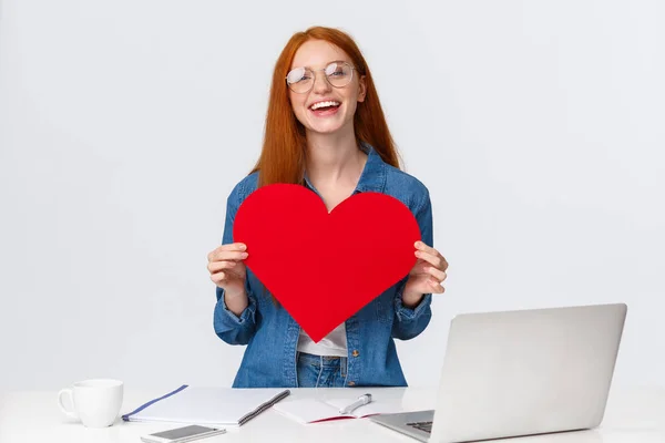 Valentines day, creativity and feelings concept. Cheerful smiling redhead girl with long-distance relatioship sending her love through internet, using web cam to show big red heart and say love you