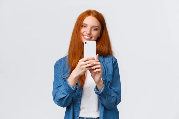 Waist-up portrait cheerful modern cute redhead woman taking photo of friend, smiling and tilt head lovely, looking mobile display, shooting beautiful scenery to post online in blog, white background