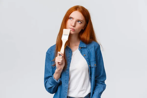 Thoughtful and creative redhead female imaging what colour paint room, holding paintbrush, look up thoughtful, thinking choices, pondering ideas, standing white background unsure