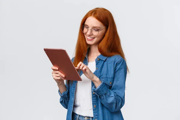 Ritratto in vita spensierata donna europea con i capelli rossi negli occhiali, guardando intrattenuto e divertito al tablet digitale, sorridente felice, soddisfatto del risultato, in piedi sfondo bianco — Foto Stock