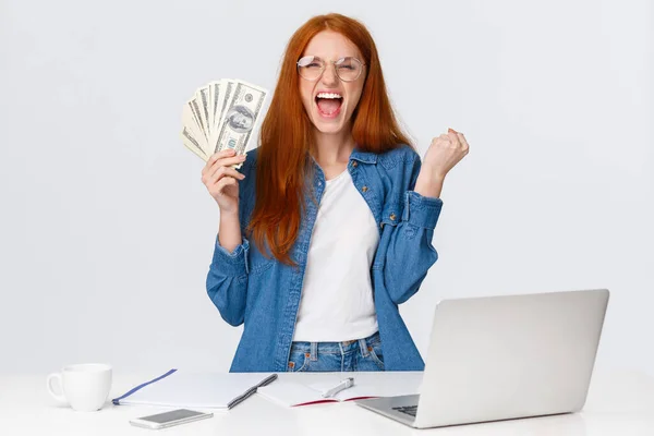 Triunfando y alegre, atractiva pelirroja feliz gritando sí, hurra con mucho dinero, mucho dinero en efectivo, tiene cheque de pago para diseñar el proyecto, recibir el pago y la celebración, fondo blanco — Foto de Stock