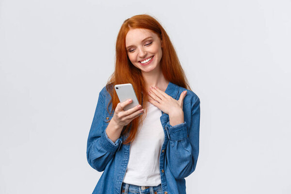 Oh so cute. Touched and pleased alluring redhead girlfriend receive love emoji, lovely message, texting, chatting boyfriend, holding smartphone, touch chest and smiling mobile display