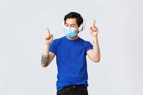 Covid-19 livsstil, människor känslor och fritid på karantän koncept. Sorglös snygg asiatisk hipster i medicinsk mask, dansar till favoritsång i hörlurar, lyssnar musik — Stockfoto