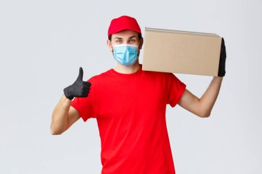 Paketler ve paket teslimatı, karantina teslimatı, nakil emirleri. Kırmızı üniformalı neşeli kurye, eldivenler ve yüz maskesi, baş parmak yukarı, müşterilere sipariş kutusu teslimatı sorun değil.