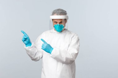 Covid-19, virüsü önleme, sağlık çalışanları ve aşı konsepti. Kişisel koruma ekipmanları içinde ciddi bir doktor. Sol üst köşeyi işaret ediyor. Bilgi panosuna.