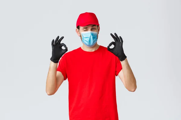 Covid-19, self-quarantine, online shopping and shipping concept. Handsome delivery guy in red cap, t-shirt, wear protective face mask and gloves from coronavirus, show okay, agree and good gesture — Stock Photo, Image