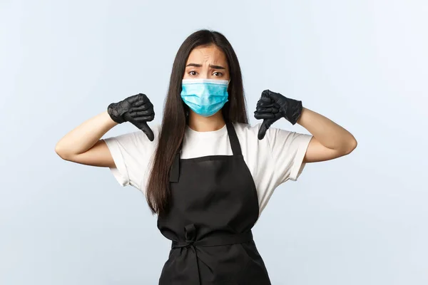 Covid-19, social distancing, small coffee shop business and preventing virus concept. Skeptical asian cafe employee in medical mask, female barista show thumb-down, dislike or disapprove