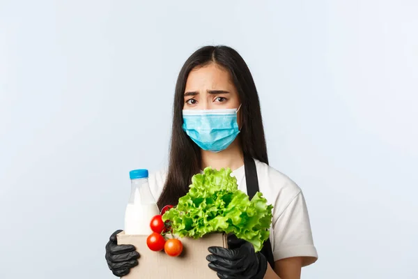 Covid-19, supermarkt, werkgelegenheid, kleine bedrijven en het voorkomen van virus concept. Bezorgd en boos fronsen aziatische vrouw winkel werknemer, kassier in medisch masker houden boodschappentas — Stockfoto