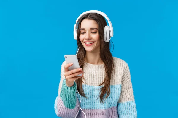 Technology, millennials and lifestyle concept. Carefree cute brunette female student put on headphones, plug-in smartphone picking song and smiling, standing blue background make playlist for study — Stock Photo, Image