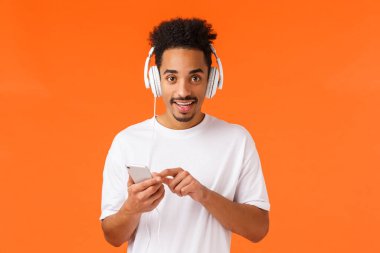 Heyecanlı, mutlu, karizmatik, Afro saçlı, bıyıklı, Afro saçlı Amerikalı hippi adam elinde akıllı telefon, step aleti seçmeli şarkı, büyük kulaklıklarla müzik dinliyor, turuncu arka plan.