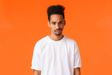 Hoşnutsuz genç Afro-Amerikan şüpheci adam, kaşlarını çatmış ve sırıtıyor, kınıyor ya da onaylamıyor, kötü fikirlerden emin değil, tereddüt ediyor plan gibi, turuncu arka plan