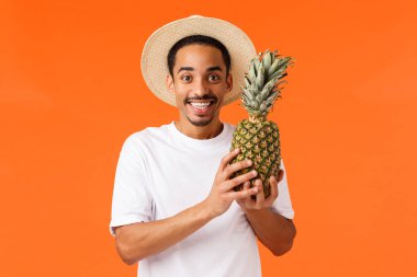 Tatilin tadını çıkarma zamanı. Mutlu gülümseyen, Afro-Amerikan erkek turist, ananas tutan ve gülümseyen erkek arkadaş, yurtdışı gezisi sırasında yerel meyveleri deniyor, lüks oteller, turuncu arka plan.