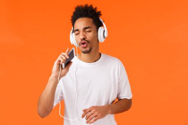 Uygulama, teknoloji ve akıllı telefon kavramı. Kaygısız Afro-Amerikan erkek karaoke oyunu oynuyor, uygulama yapıyor, mikrofona şarkı söylüyor, kulaklık takıyor, müzik dinliyor.
