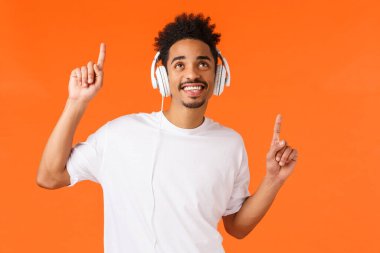 Beyaz tişörtlü mutlu Afro-Amerikan adam, yukarı bakıyor ve gülümsüyor kulaklık kullanarak müzik dinliyor, neşeli hareketler yapıyor, yeni şarkının tadını çıkarıyor, havalı müzik listesi, turuncu arka plan