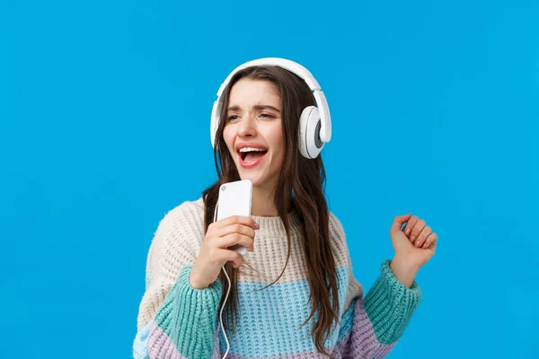 Retrato en la cintura sin preocupaciones guapa joven morena jugando karaoke juego en el teléfono, sosteniendo teléfono inteligente como micrófono, cantando en el baile del teléfono, escuchar música en los auriculares, fondo azul — Foto de Stock